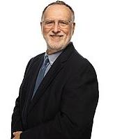 John Brunton stands in front of a white background. He is wearing a black suit and smiling toward the camera.